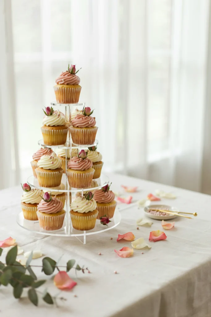 A modern clear acrylic five-tier cupcake tower holding white and blush cupcakes at different heights, surrounded by scattered rose petals on a white linen-covered table