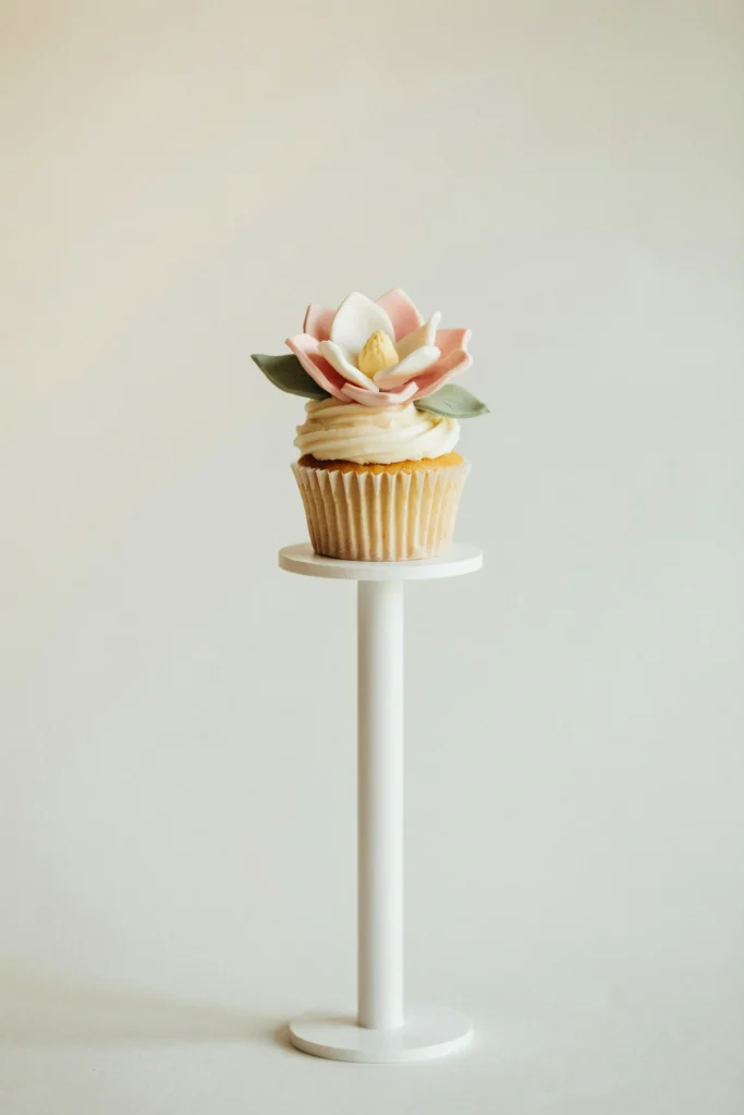 A single cupcake on an elegant white pedestal — smooth ivory cream cheese frosting topped with a handcrafted fondant magnolia bloom in blush pink and cream, with two small fondant leaves