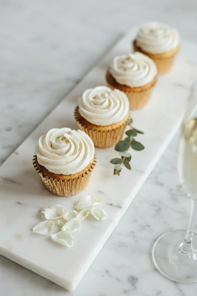 Close-up of a wedding cupcake topped with a perfectly piped large white buttercream rose, sitting on a gold foil liner, surrounded by three similar cupcakes on a marble surface