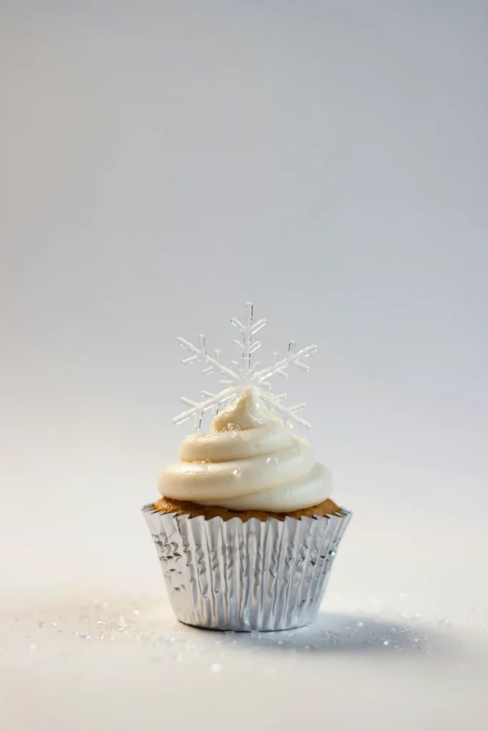 A pure white cupcake on a silver foil liner with smooth pure white frosting, a single crystallized isomalt snowflake balanced on top — glassy, translucent, catching overhead light like ice