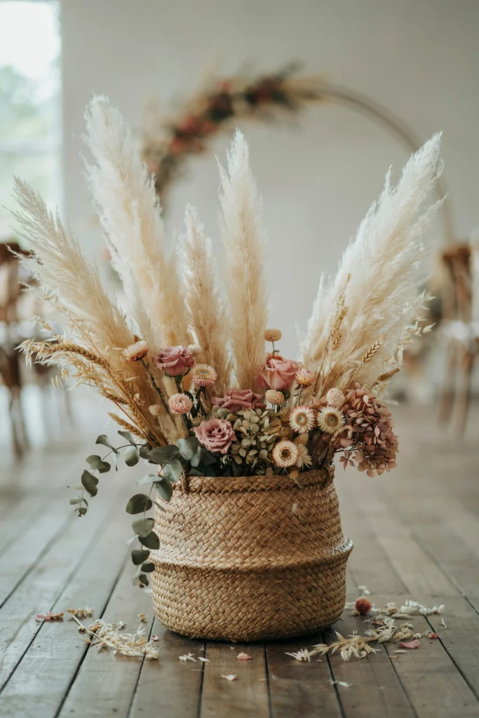 A large woven basket overflowing with dried wheat, pampas, and dusty pink dried flowers, placed at the base of the floral arch or as a freestanding floor arrangement near the entrance.