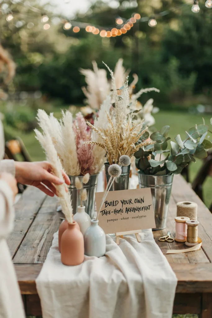 A styled station with buckets of dried grasses, pampas, and preserved eucalyptus sorted by texture, ribbon spools, and small vases, where guests can build their own take-home arrangement.