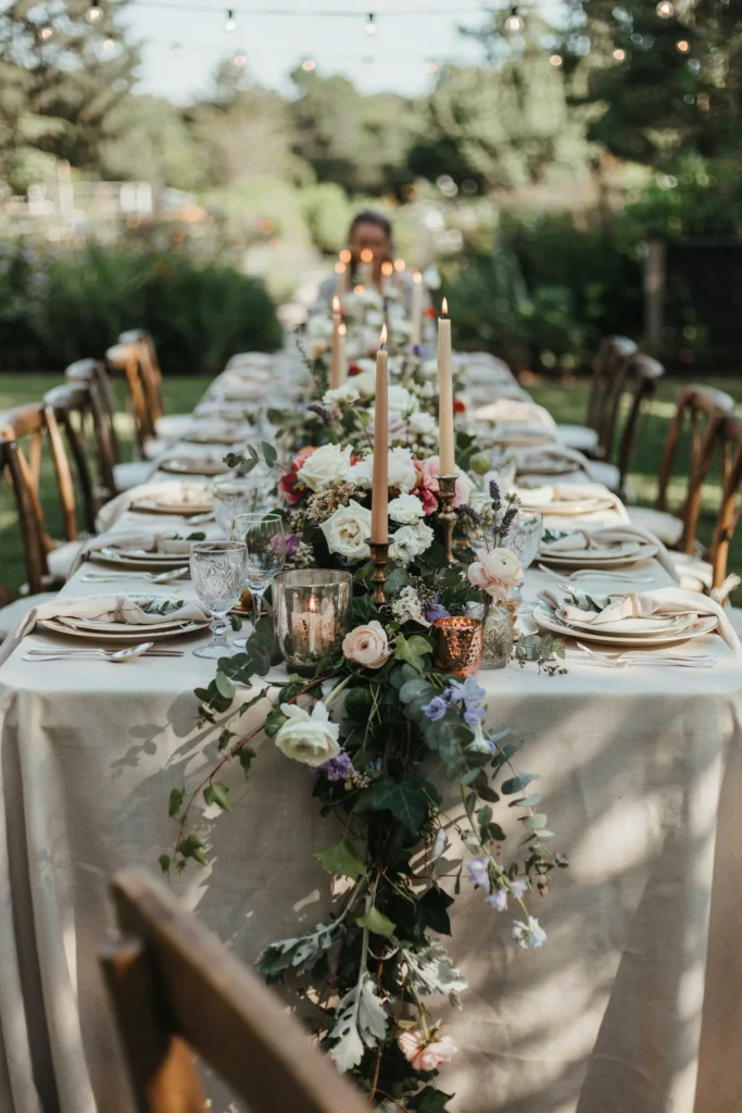 A table runner made entirely of a loose, lush floral and greenery garland cascading down the center of a long table, with mismatched candle holders nestled between the blooms.