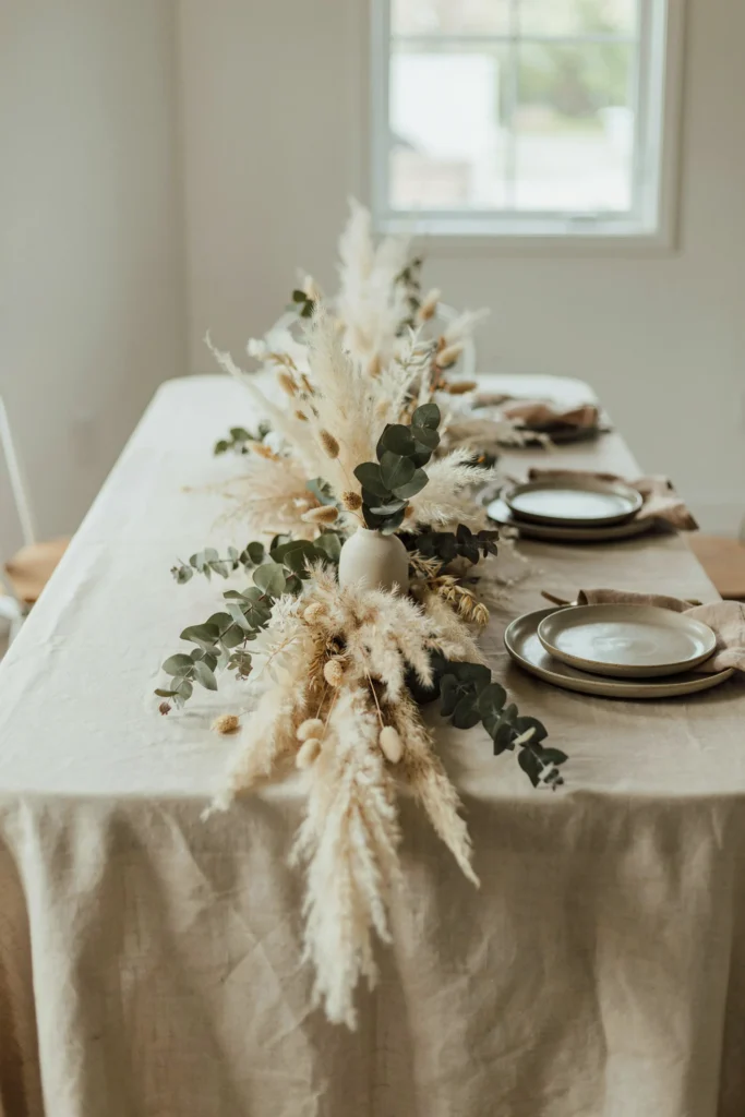 A long, loose centerpiece running down the center of a table, made entirely of dried pampas grass, preserved eucalyptus, and dried grasses in soft neutral tones, with a few stems trailing off the edge of the table.