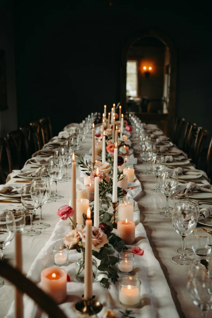 A long table lined with dozens of flameless LED candles at varying heights between low floral arrangements, the warm flicker reflecting off glassware in a dimly lit room.