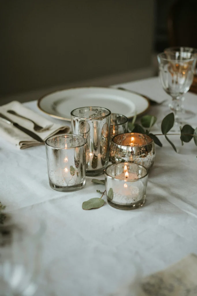 A grouping of mercury glass votive holders in varying sizes, each holding a small flameless candle, clustered at the center of a place setting or along the table's edge.