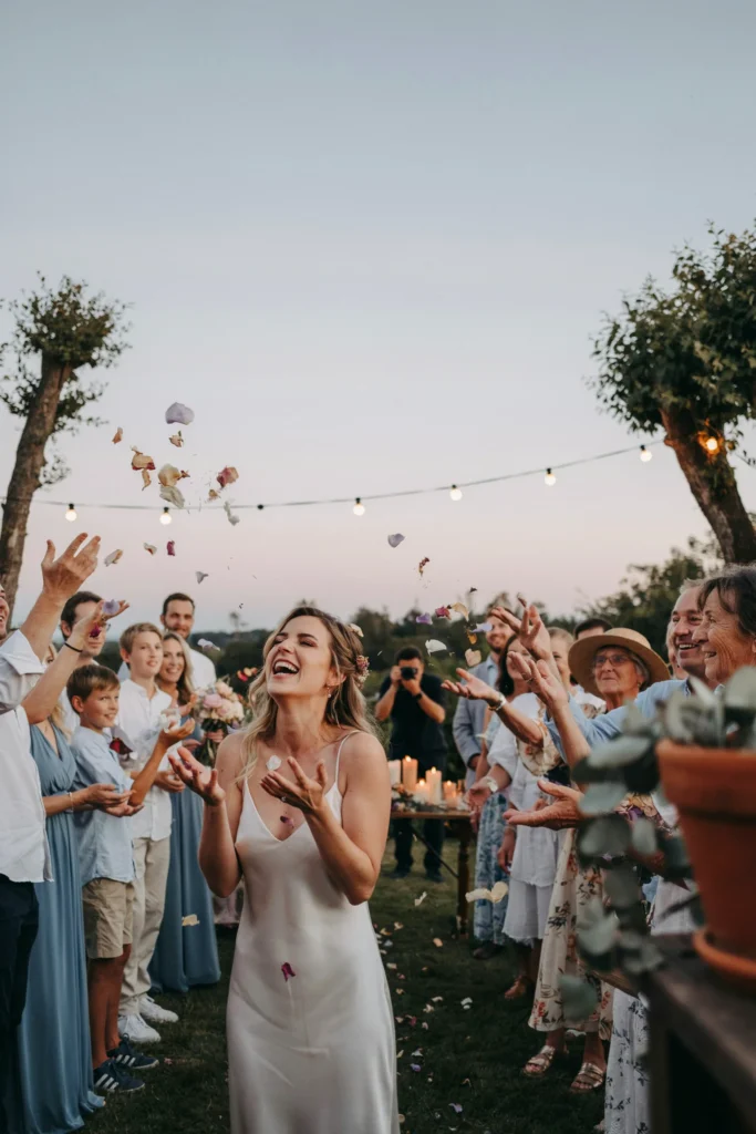 Guests gathered in a loose circle around the bride at golden hour, dried flower petals and confetti caught mid-air in a single photograph, candlelight just beginning to glow in the background as dusk settles.