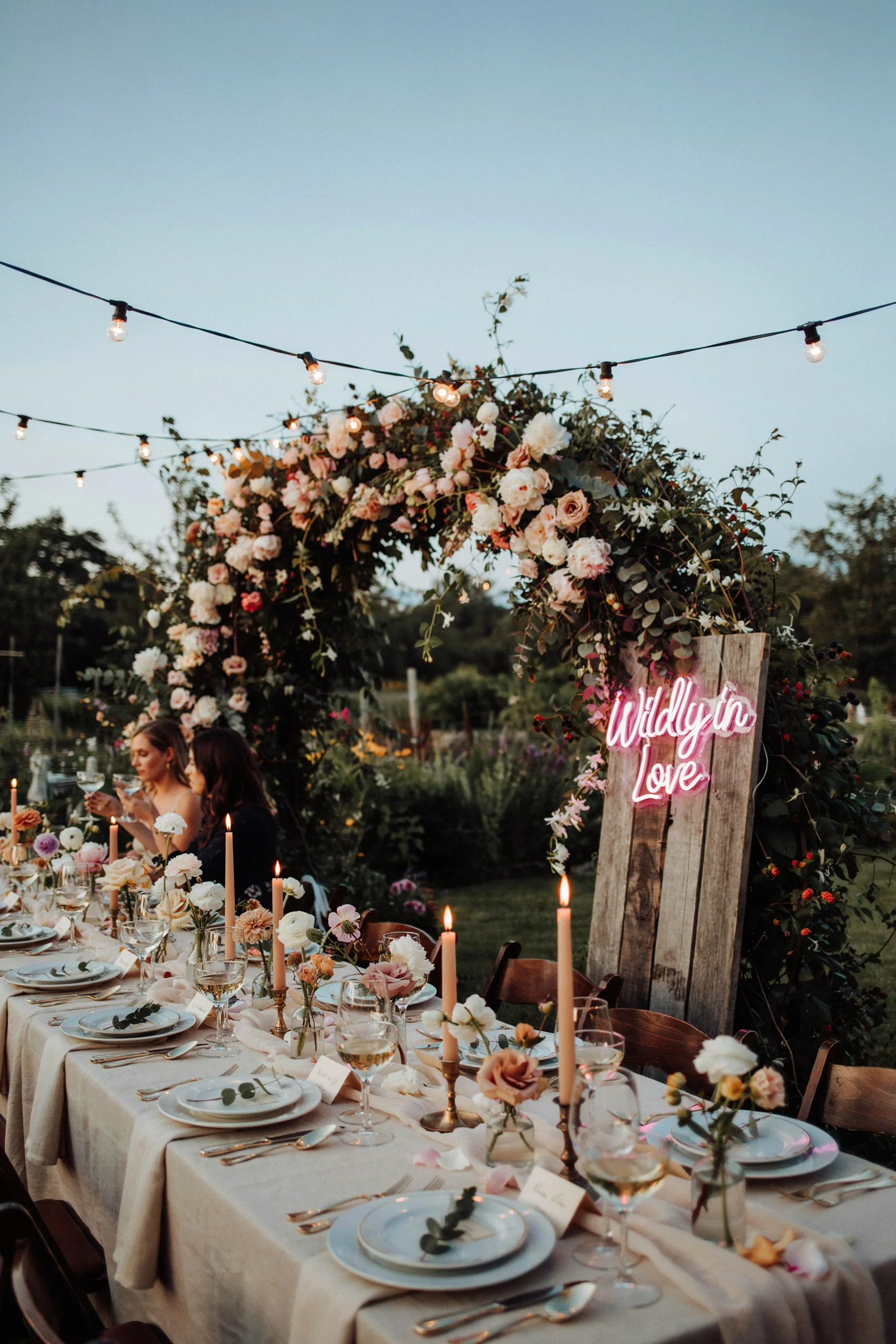 An evening bridal shower table glowing under string lights and an oversized floral arch, candlelight reflecting off champagne coupes and a "Wildly in Love" neon sign in the background. Mood: lush, romantic, slightly untamed