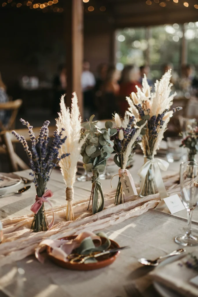 Small bundles of dried lavender, pampas, and eucalyptus tied with ribbon, displayed in a row on a table for guests to take home, each bundle slightly different in composition.
