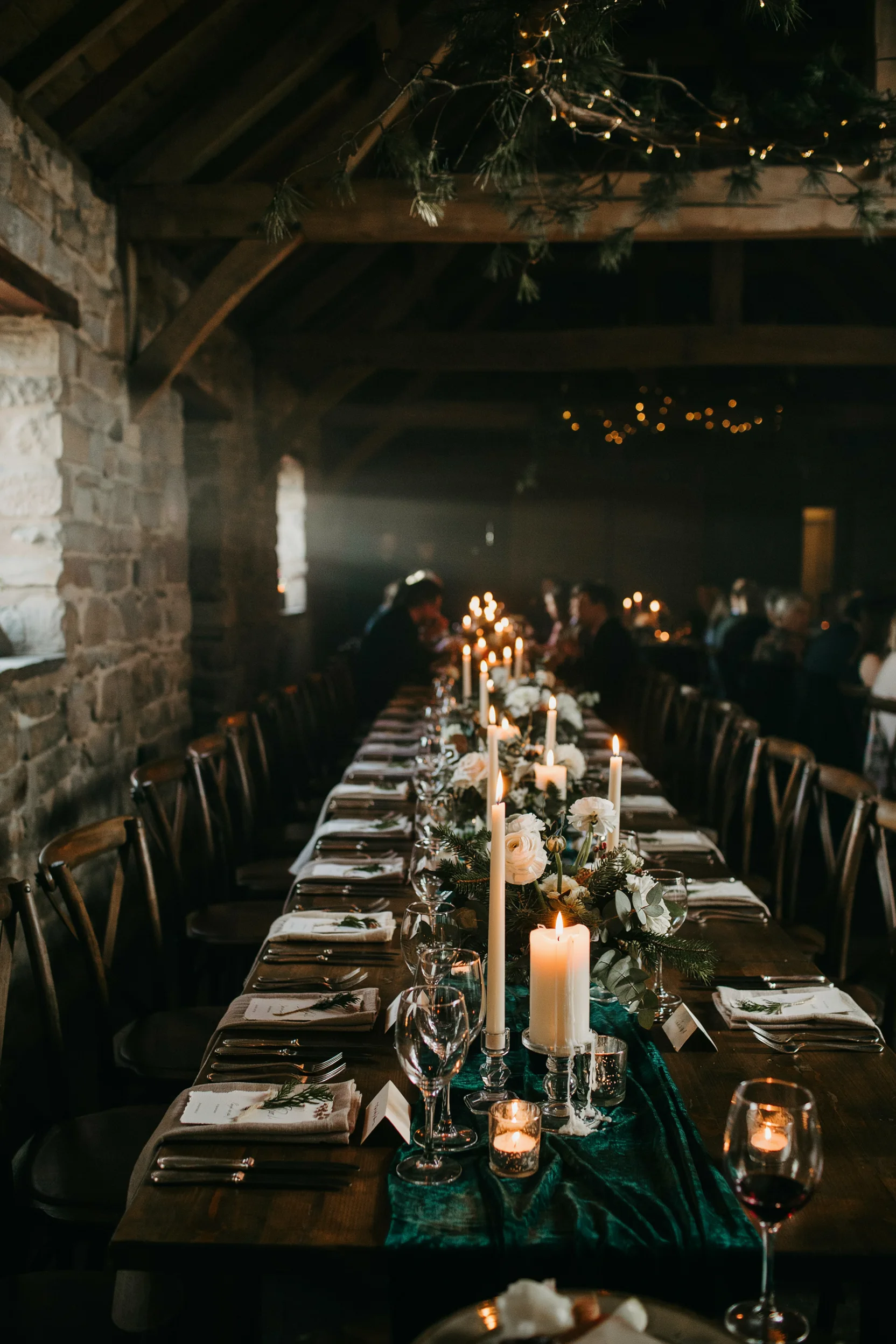 A moody, candlelit winter wedding reception with long tables draped in deep emerald velvet runners, towering pillar candles, white ranunculus and pine branch centerpieces, and warm amber lighting.