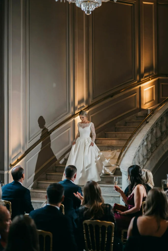 A bride descending a wide marble staircase in a full ballgown, guests below looking up, dramatic uplighting on the walls