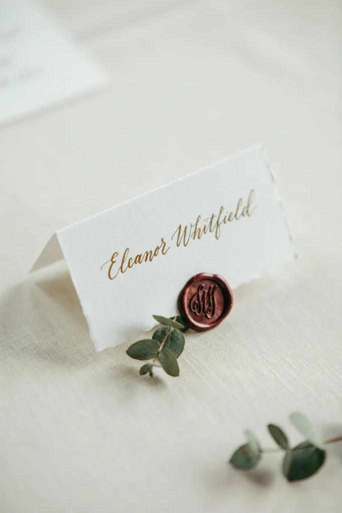 Ivory place cards with gold calligraphy names, a burgundy wax seal, and a small sprig of greenery tucked underneath