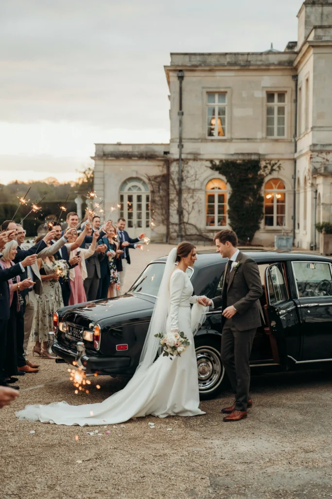 A vintage black Rolls-Royce parked outside a formal venue at golden hour, bride and groom stepping in, guests waving with sparklers
