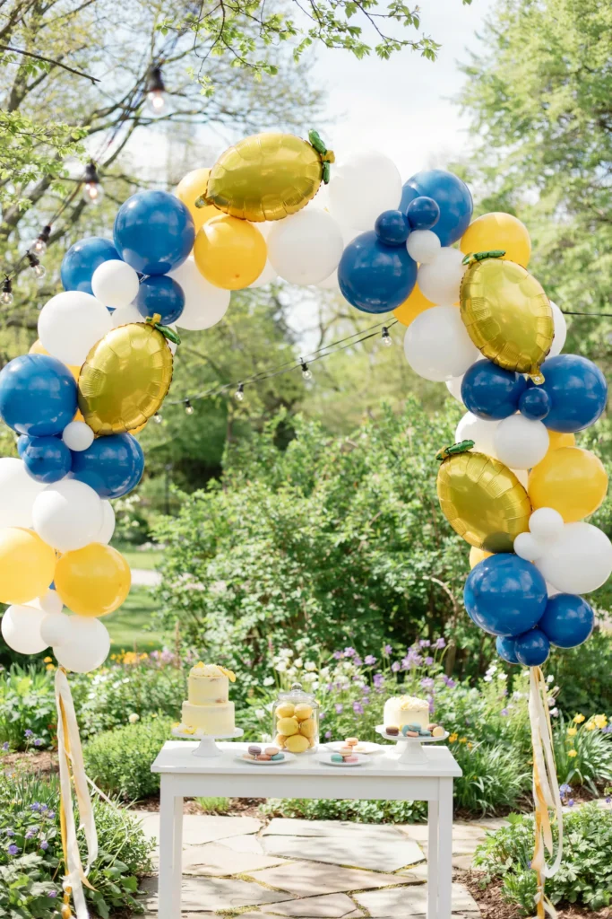 Organic balloon arch in royal blue, lemon yellow, and white framing a doorway or dessert table, with a few oversized lemon-shaped balloons tucked into the cluster.