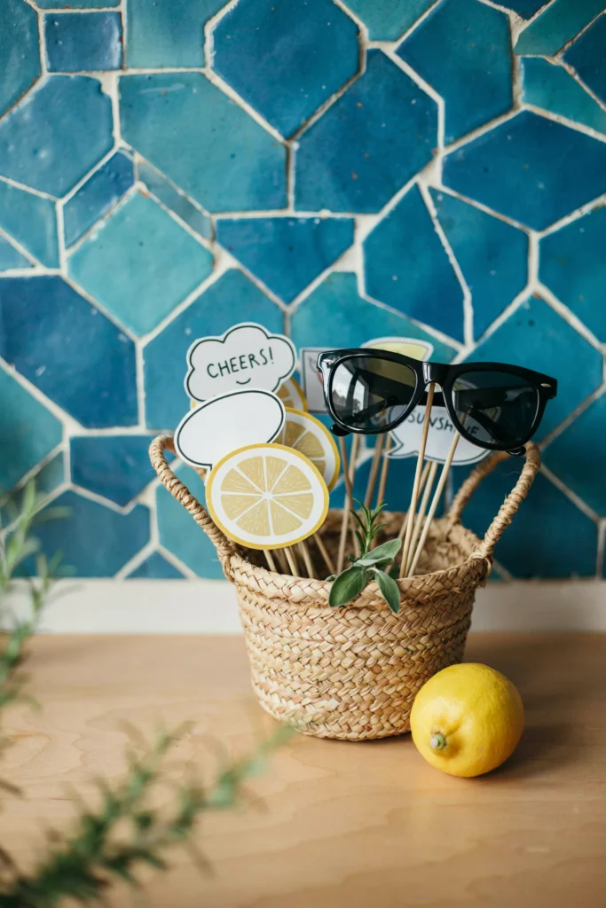 A small styled corner with a blue-tile backdrop and a basket of lemon and citrus-themed photo booth props on sticks — sunglasses, lemon slices, speech bubbles.