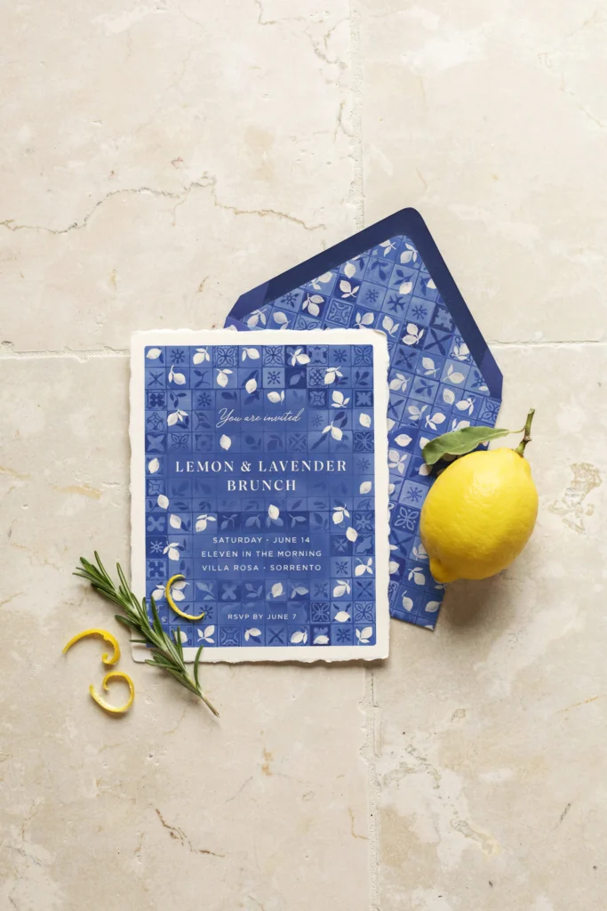 Flat-lay of a blue-tile lemon-print invitation with matching envelope, styled next to a lemon and a small sprig of greenery.