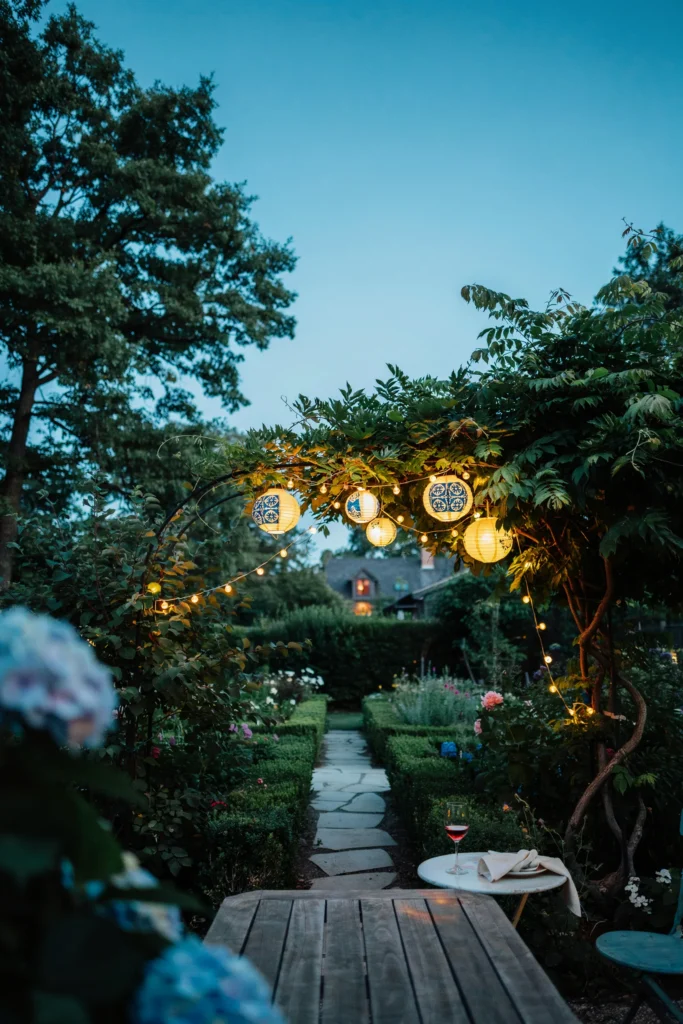 Outdoor garden setting at dusk with hanging paper lanterns in yellow and blue tile patterns, string lights woven between trees