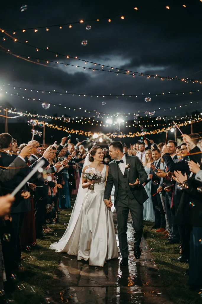 Nighttime wedding send-off with guests blowing gold-tinted bubbles under string lights
