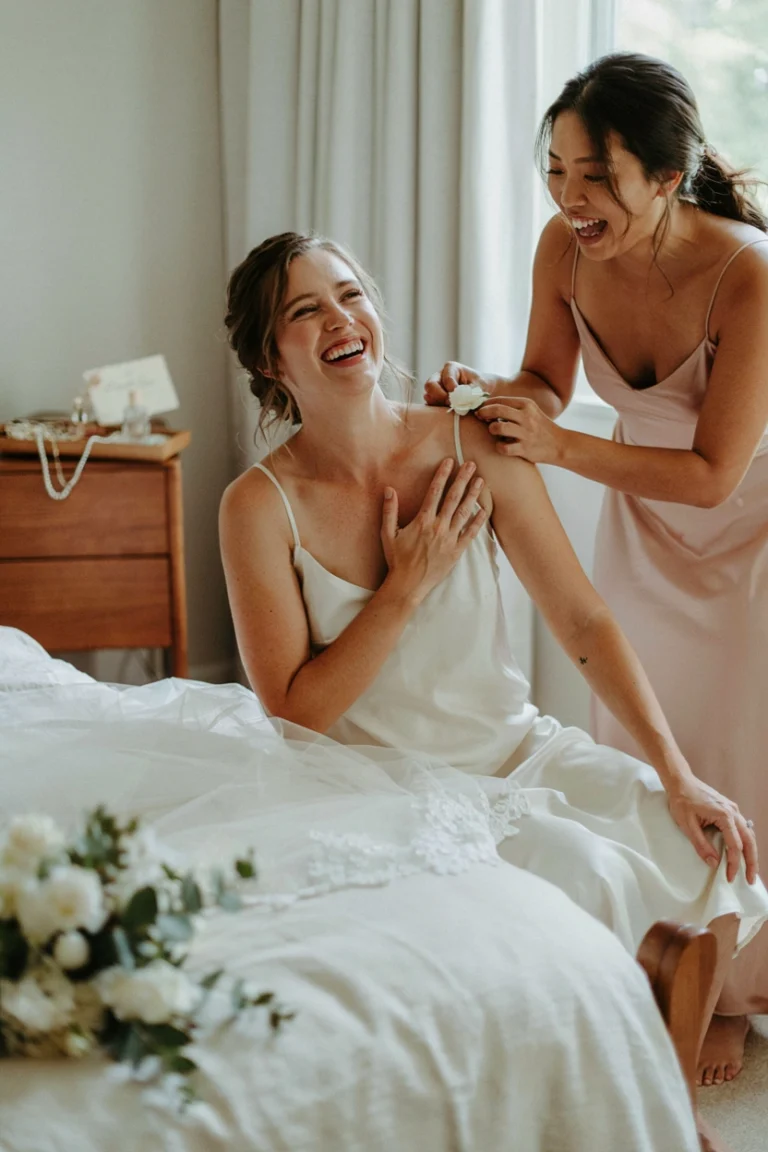 A maid of honor helping a bride get ready, laughing together while pinning a boutonniere or adjusting a veil — warm, candid, natural light.
