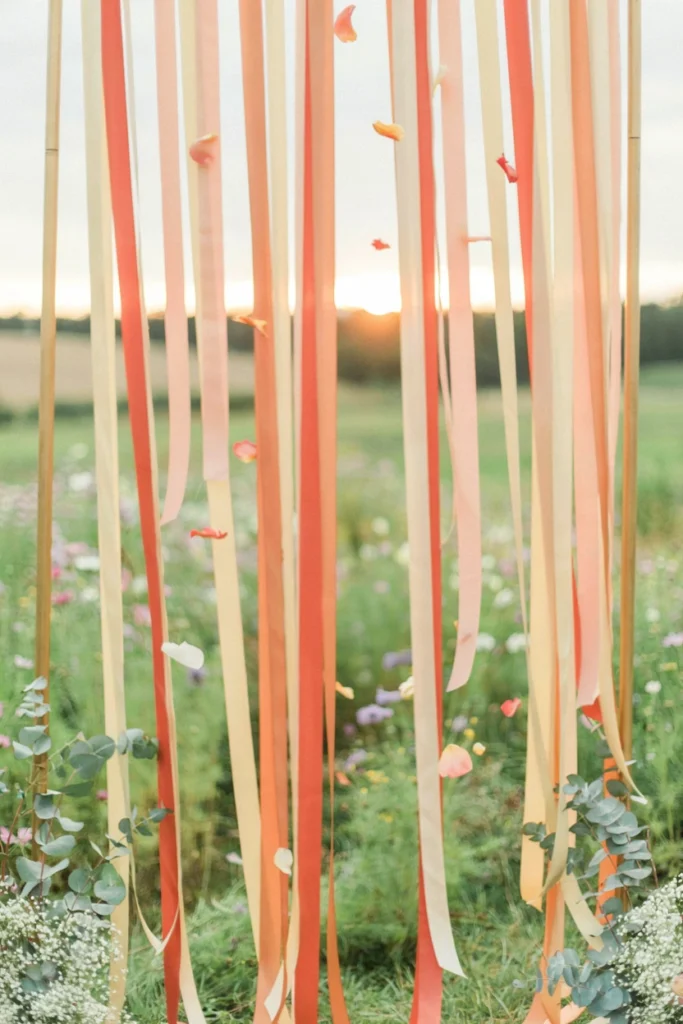 A backdrop made of flowing ribbon or fabric streamers in a gradient from deep coral at the top to pale gold at the bottom, set up facing west so the actual sunset lights it from behind