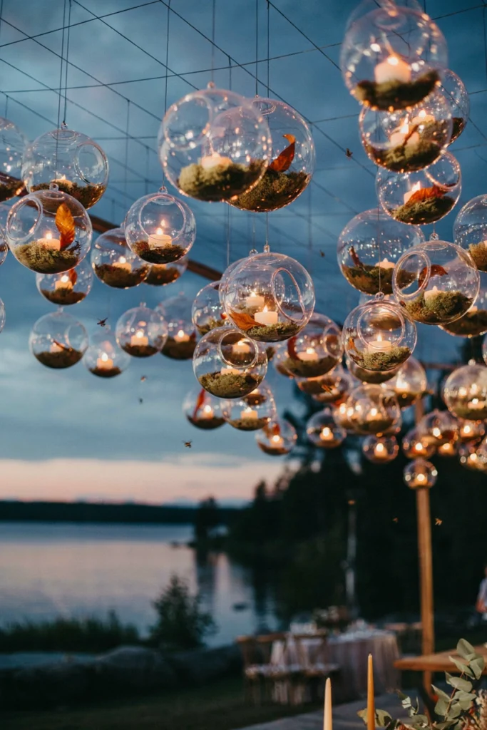 Hanging glass terrarium candles with moss glowing above a lakeside wedding reception at dusk