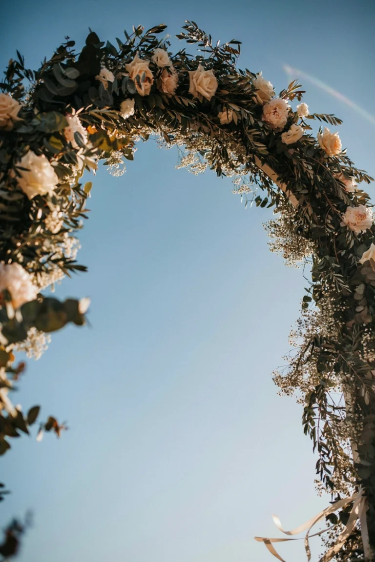 A ceremony arch wrapped in a lush eucalyptus and white rose garland, shot from below so the greenery frames a clear blue sky, soft golden-hour light catching the edges of the leaves. Mood: romantic, lush, aspirational.