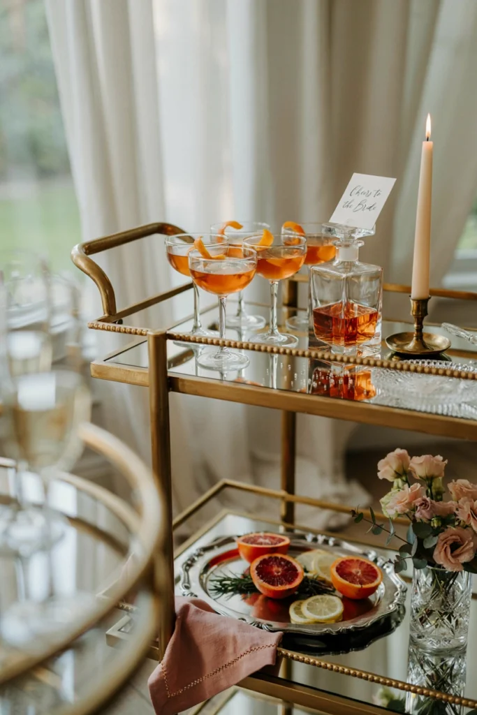 A small bar cart or table with amber and pink cocktails in clear glassware, citrus garnish, and a reflective metallic tray catching candlelight