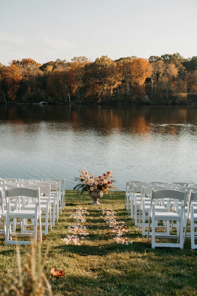 White folding chairs lining a minimalist petal aisle ending at the lake's edge with a single low floral urn