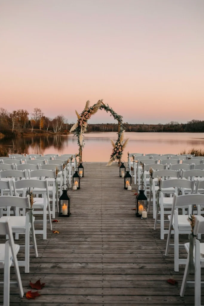 Rows of ceremony chairs on a wooden dock lined with glowing black lanterns at sunset