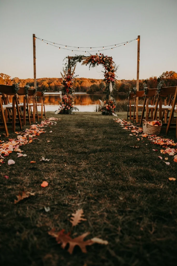Grass wedding aisle scattered with peach and rust rose petals leading to a floral arch at sunset by a lake