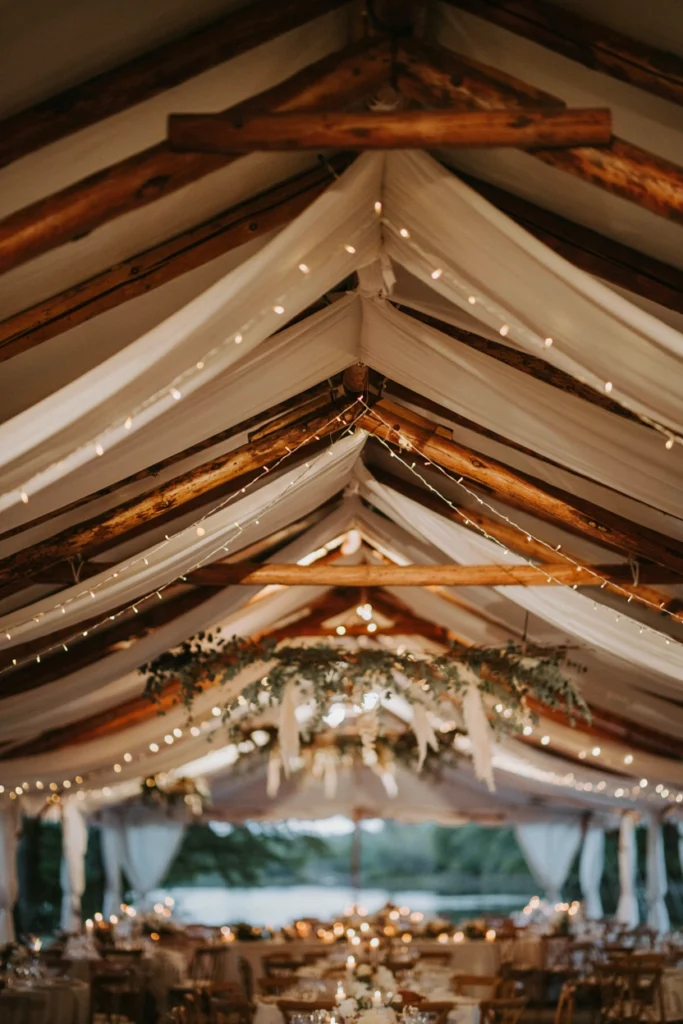 Draped white fabric ceiling with warm amber string lights over a lakeside wedding reception tent