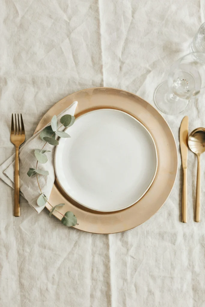 Overhead place setting with a champagne metallic charger plate, gold flatware, and a sprig of eucalyptus