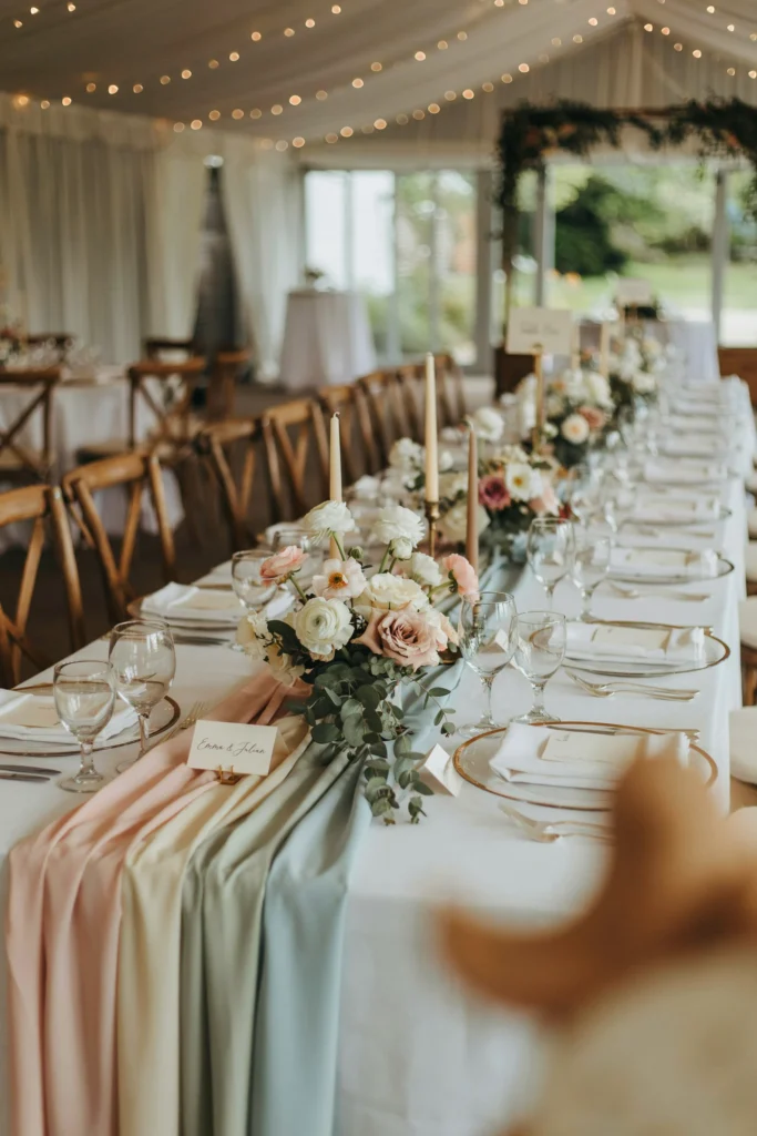 A long reception table with alternating pastel satin runners crossing at angles, catching light with a soft sheen against white linens.