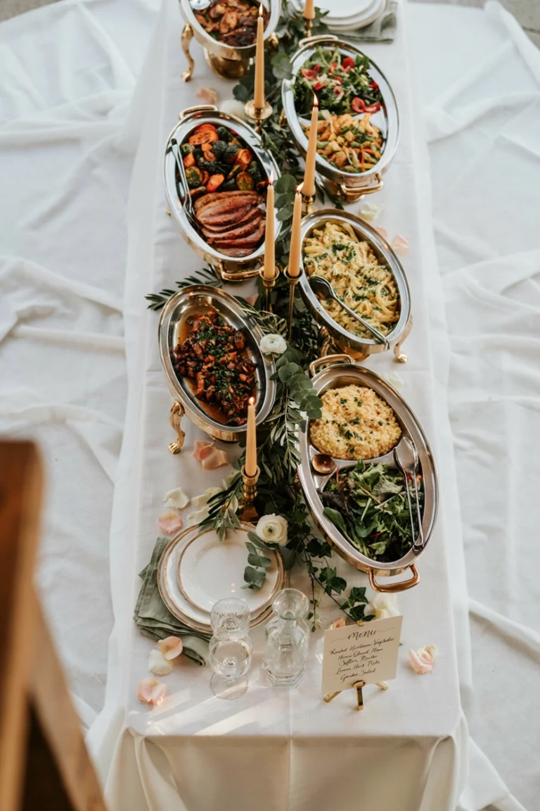 An overhead shot of a beautifully styled wedding buffet table with white linens, brass chafing dishes, greenery runners, and soft golden light — warm, abundant, and celebratory.