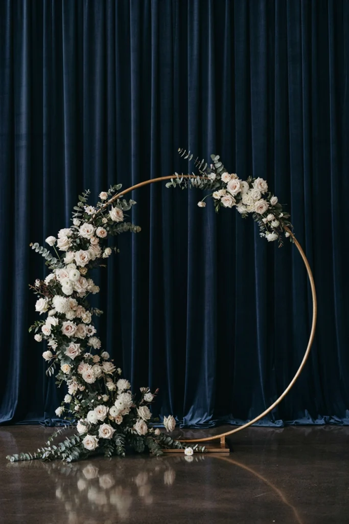 Gold circle wedding arch with cascading white floral arrangement against navy drapery
