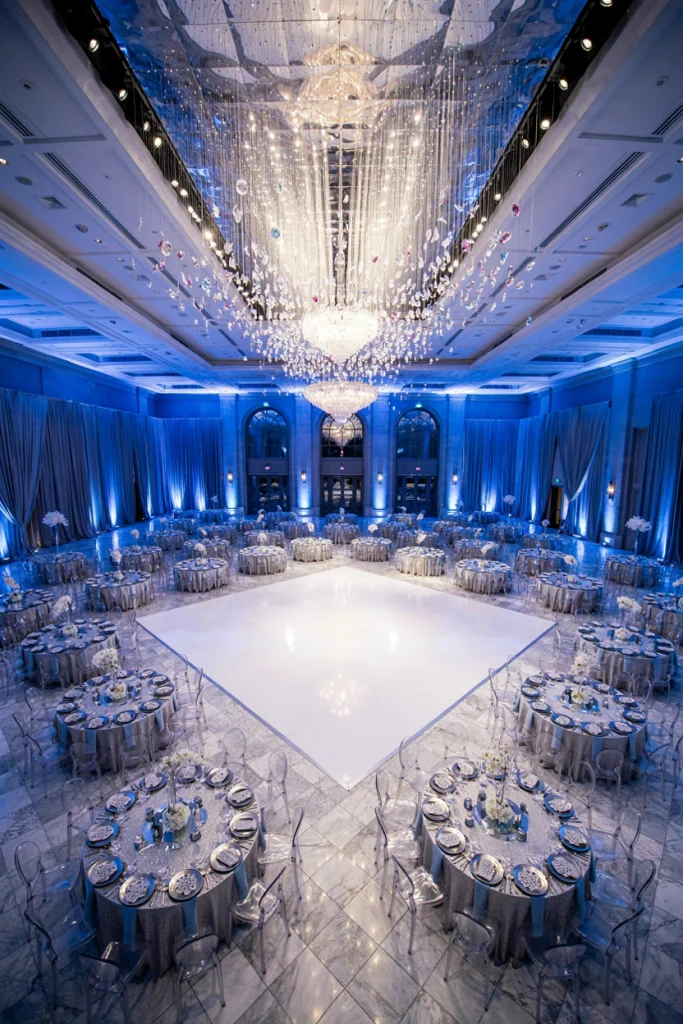 Large icy blue and silver ballroom wedding reception with rows of round tables and blue uplighting